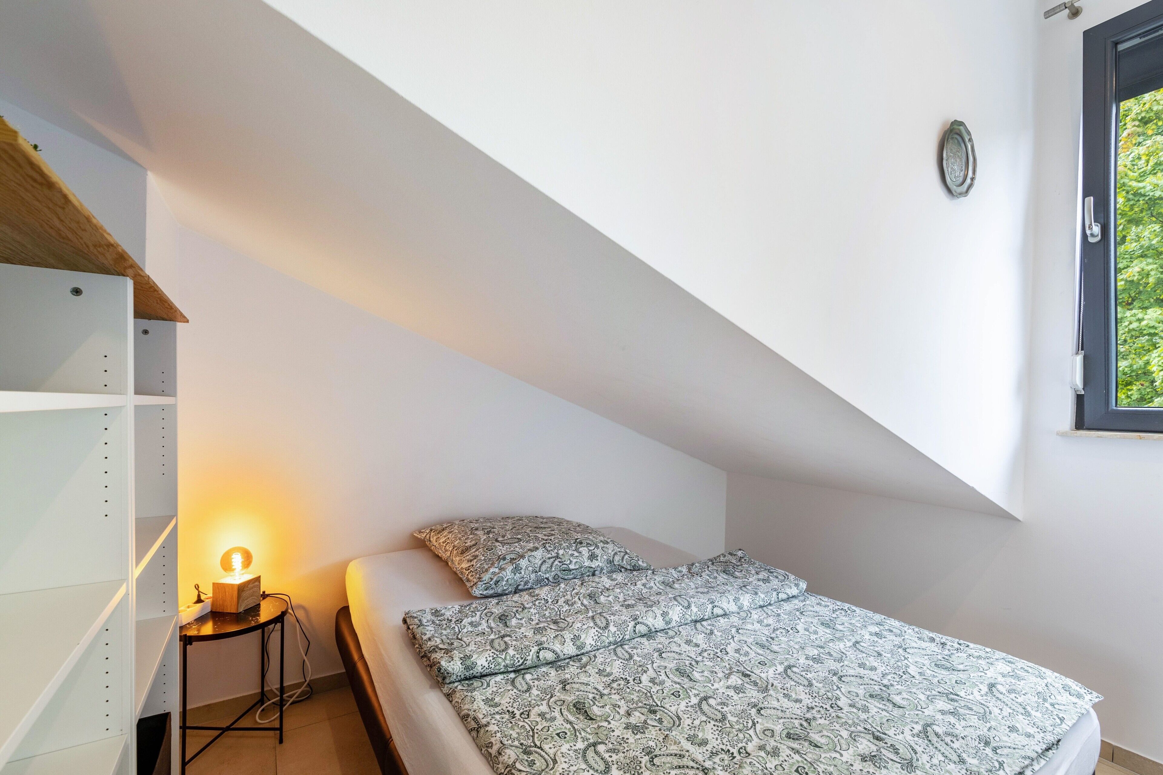 A bedroom with a sloped ceiling, a bed with patterned bedding, a bedside table with a lamp, and a window with a view of greenery.