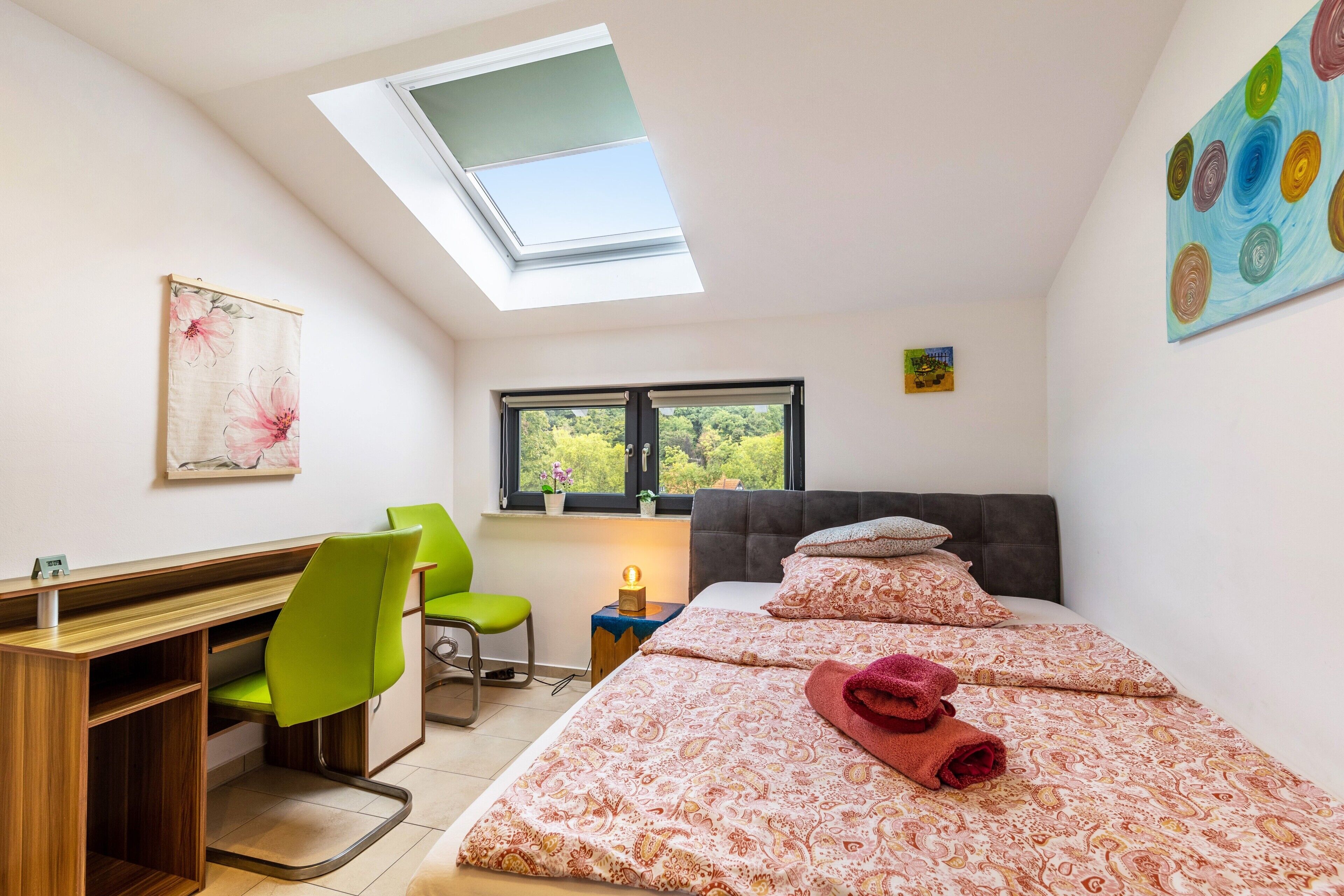 A bedroom with a skylight, a desk, a chair, a bed with patterned bedding, and a window with a view of greenery.