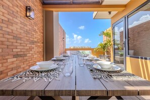 A wooden dining table set with white plates, bowls, and glasses, with a brick wall and a glass door on one side.