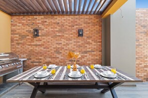 Un espace extérieur aménagé pour les repas, avec un mur en briques en arrière-plan, un barbecue et une table dressée pour quatre personnes.