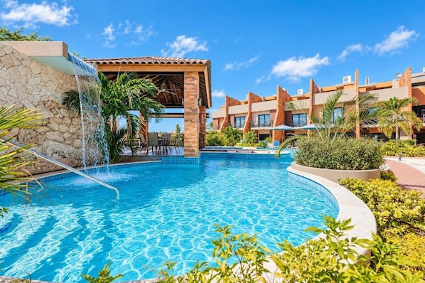 A swimming pool with a waterfall feature, surrounded by a poolside area with chairs and a covered seating area.