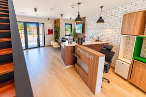 A reception desk with a wooden finish and the name 'BOCOBAY' on it. There is a seating area with chairs and a potted plant. The room has a brick wall and a large window.
