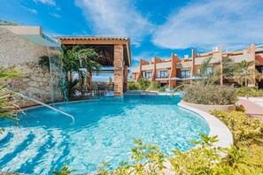 A swimming pool with a waterfall feature, surrounded by a building with multiple balconies and a clear blue sky. - LUX 3BR House Pool Gym 7-Min Walk to Palm Beach - Porta al Sole 19 by Bocobay (Noord)