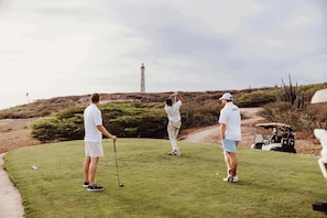 Ein Golfplatz mit Leuchtturm im Hintergrund, einem Golfwagen und Spielern in weißer Kleidung.