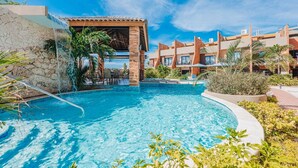 Une piscine agrémentée d’une cascade, entourée d’un bâtiment doté de nombreux balcons, le tout sous un ciel d’un bleu limpide.