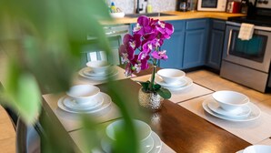 Une table à manger dressée avec de l’assiette et des bols blancs, un vase rempli de fleurs violettes, et une cuisine en arrière-plan.