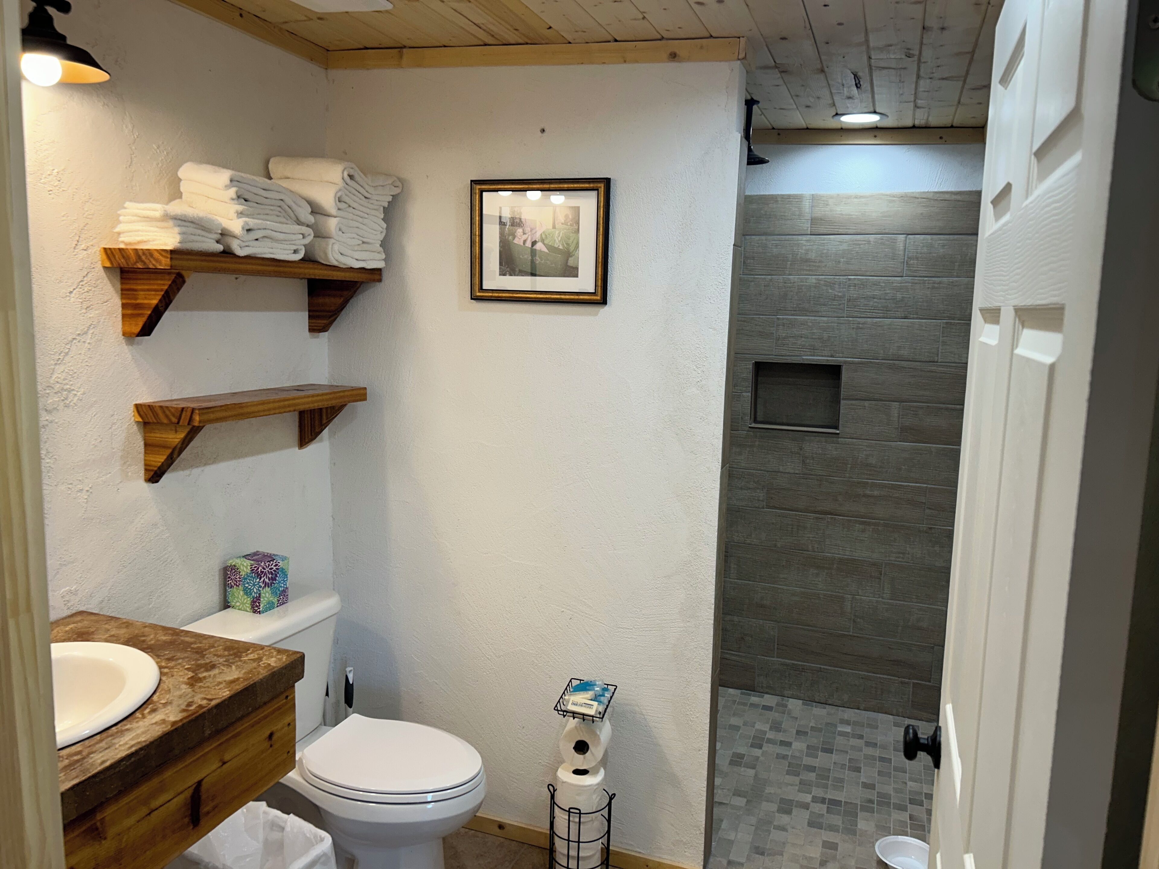Un baño con inodoro, lavamanos y zona de ducha. Hay estantes de madera con toallas y un cuadro enmarcado en la pared.