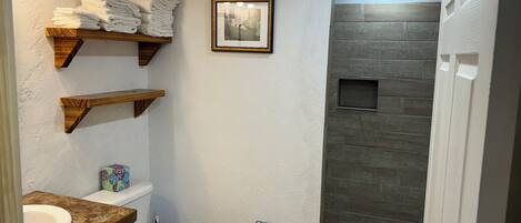 Un baño con inodoro, lavamanos y zona de ducha. Hay estantes de madera con toallas y un cuadro enmarcado en la pared.