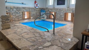 Piscina interior com soalho em pedra, escada de metal, paredes de pedra, janelas de madeira e um claraboia.