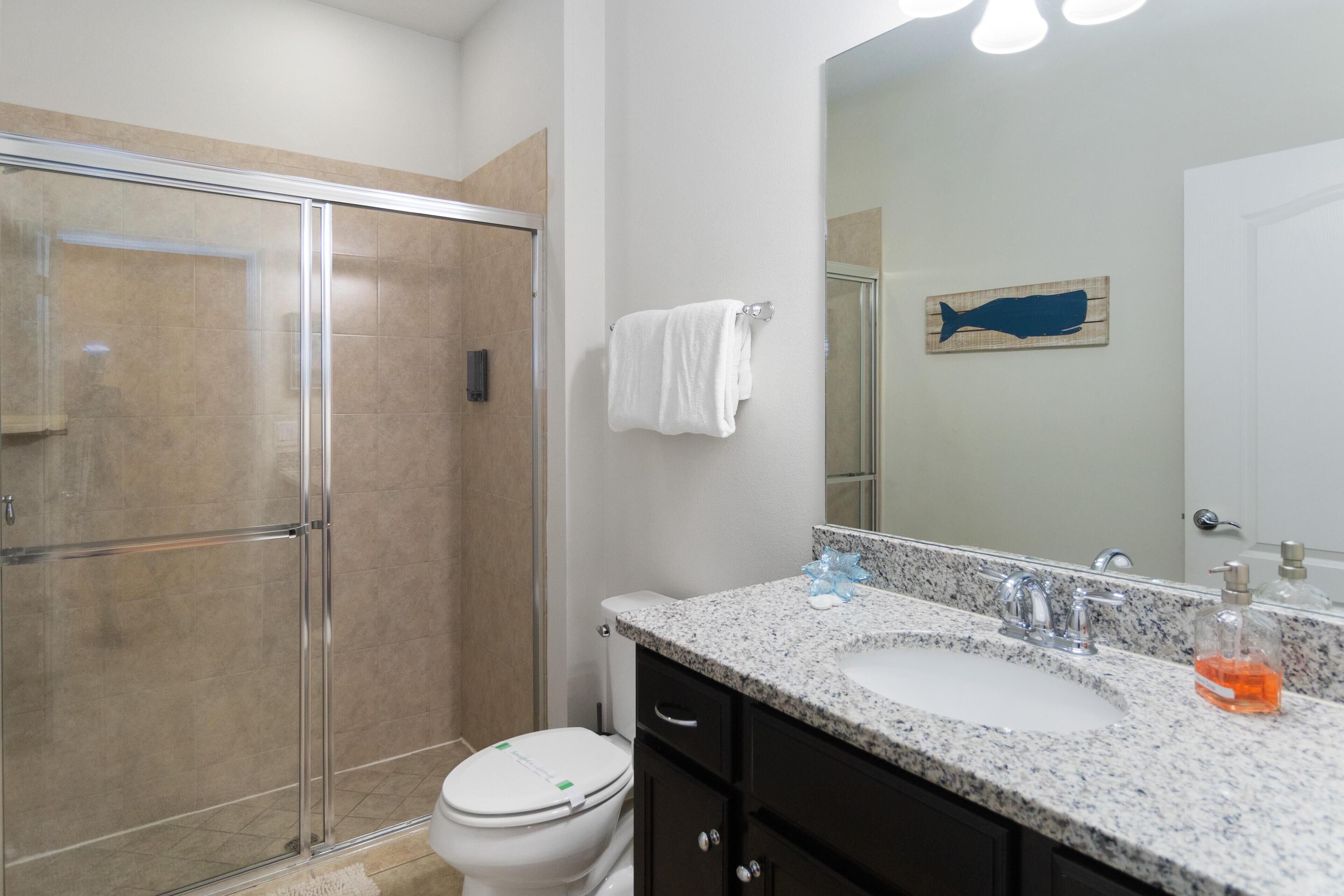 A bathroom with a glass shower enclosure, a white toilet, a granite countertop sink, and a mirror with a whale decal.