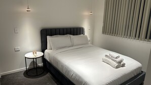 Un lit bien fait, avec du linge de lit blanc et des serviettes pliées, une petite table ronde avec une lampe, et une fenêtre avec des stores.