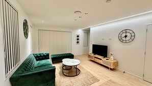 Un salon moderne avec un canapé vert, une table basse ronde, un téléviseur posé sur un meuble en bois et une horloge au mur.