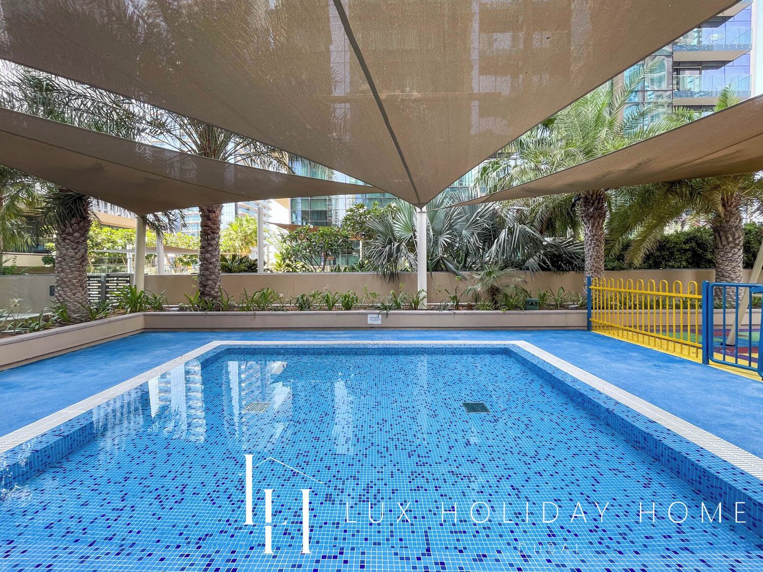 A swimming pool with a tiled floor, surrounded by palm trees and a building in the background.