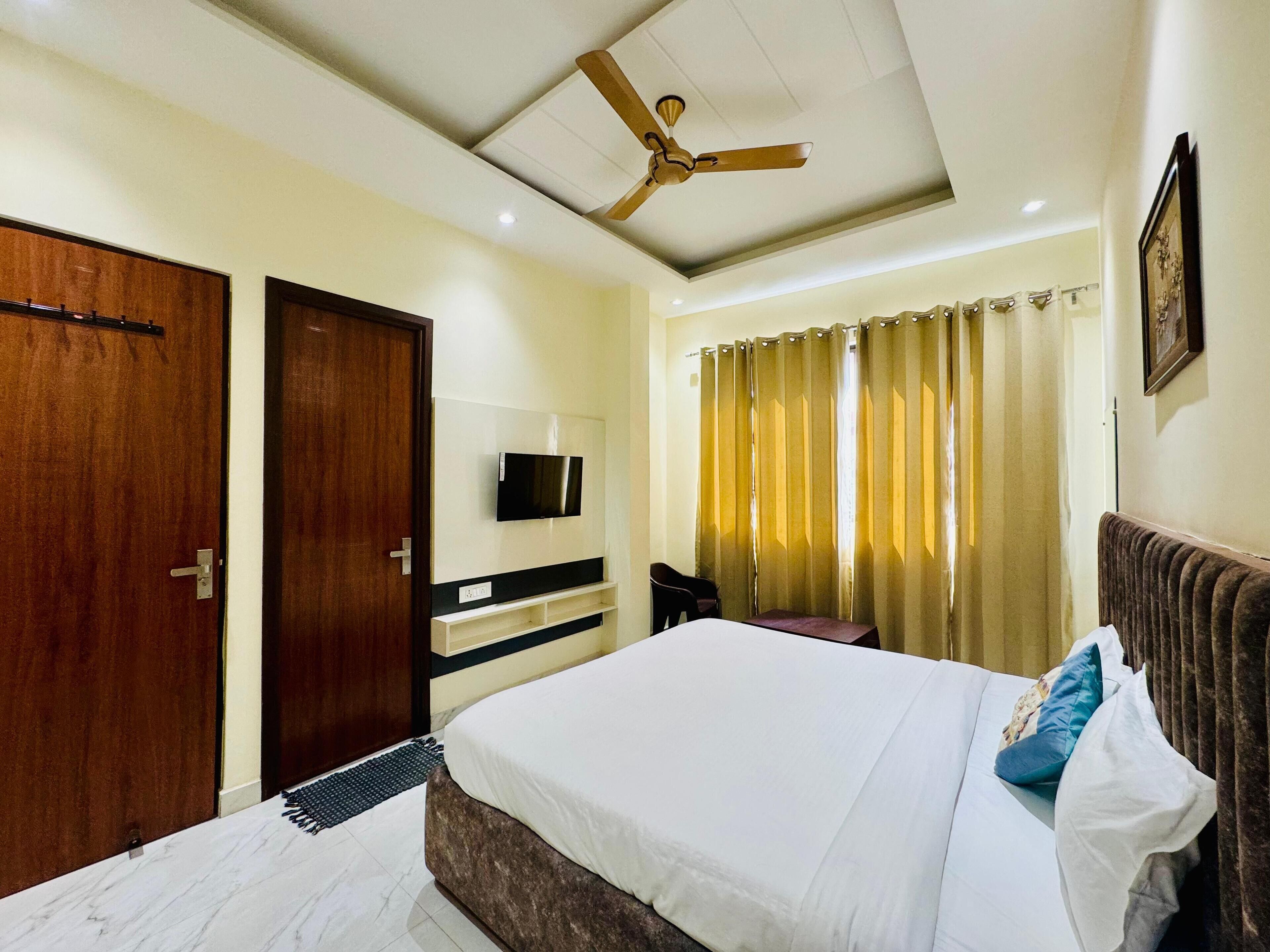 Habitación de hotel con cama, ventilador de techo, televisor montado en la pared y ventana con cortinas.