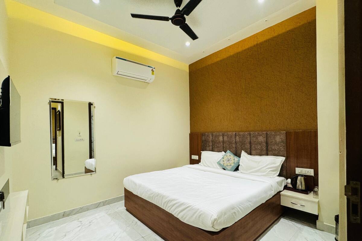 A modern hotel room with a large bed, a ceiling fan, an air conditioning unit, a mirror, and a bedside table with a lamp.