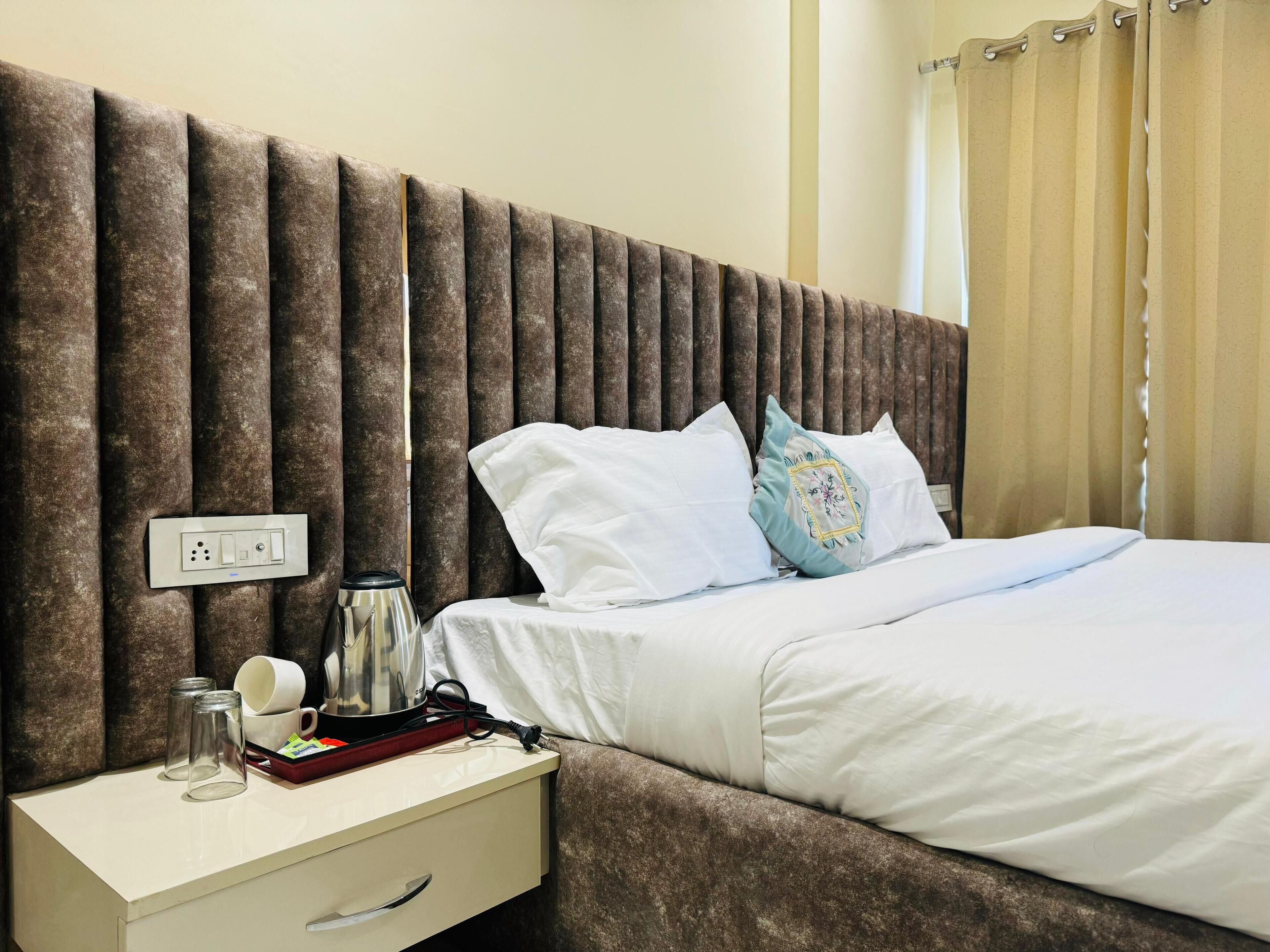 A neatly made bed with white linens and decorative pillows, a bedside table with a kettle and cups, and a wall with a textured panel design.
