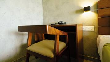 A small, compact room with a wooden desk and chair, a bedside table, and a wall-mounted light fixture.