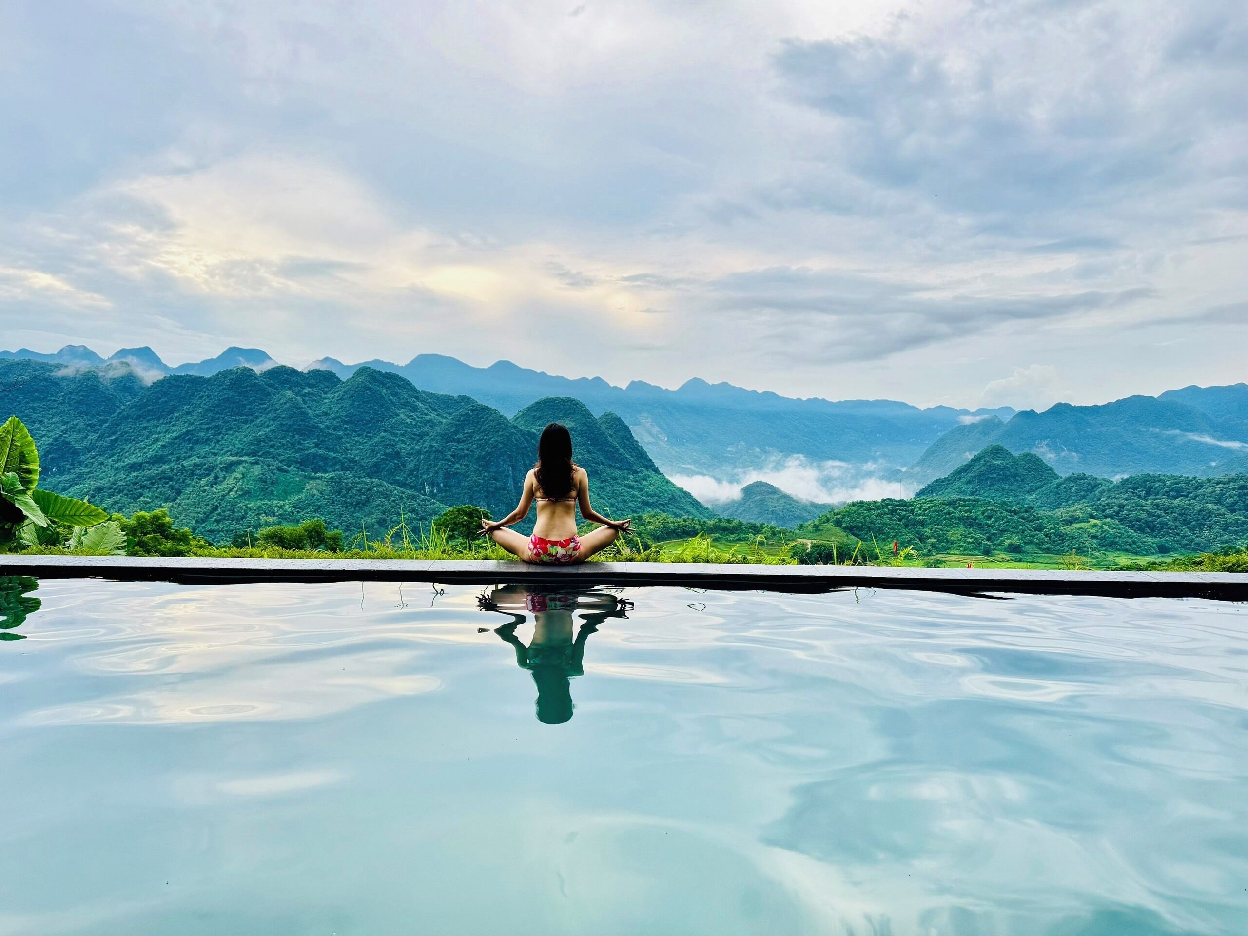 A person meditating by a reflective pool with a mountainous landscape in the background.