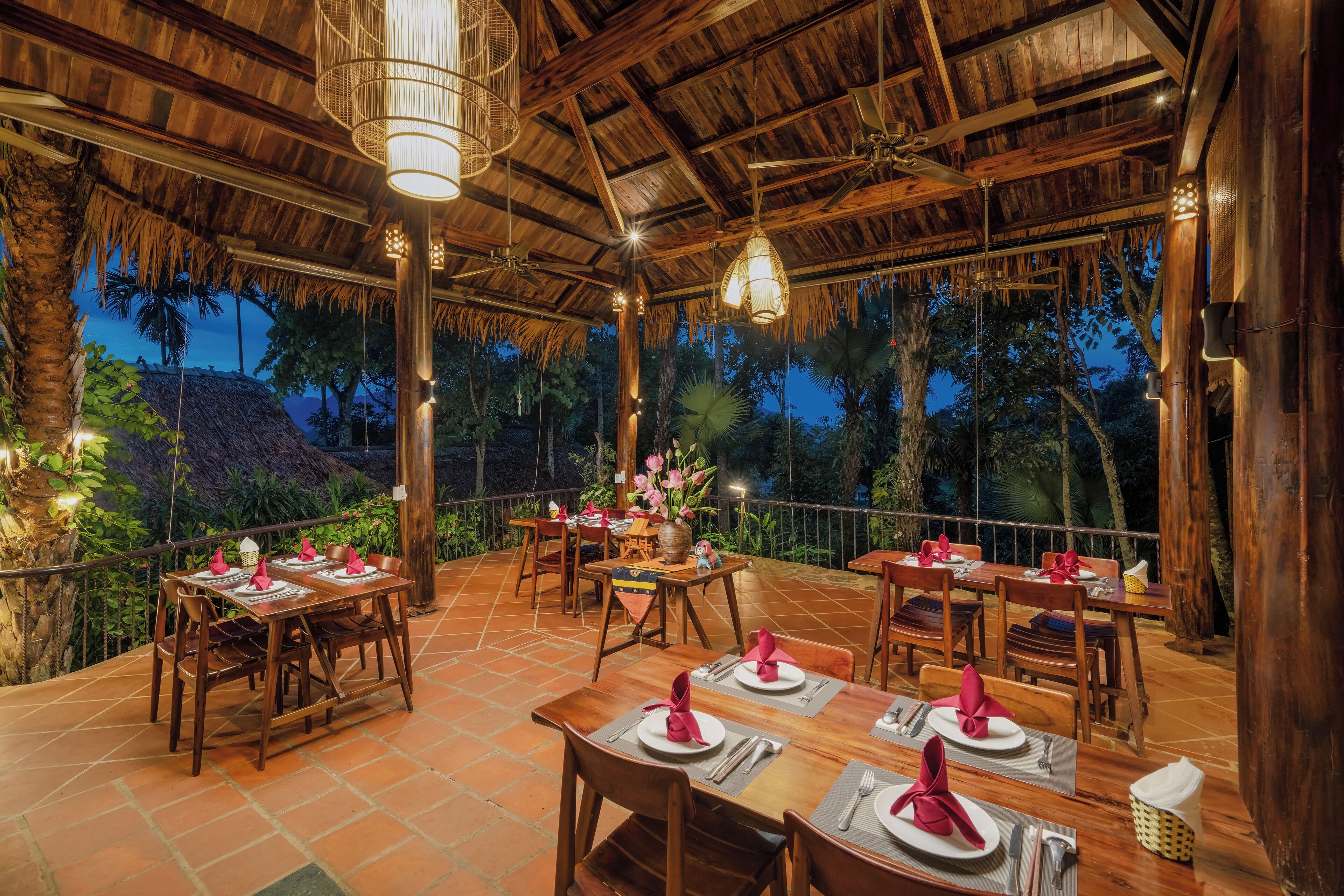 Un área de comedor al aire libre con mesas y sillas de madera, rodeada por un techo de paja y palmeras.