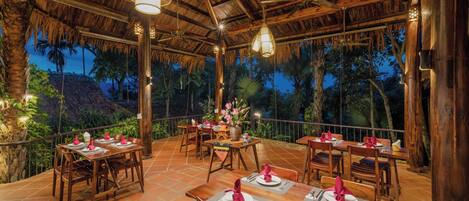 Un área de comedor al aire libre con mesas y sillas de madera, rodeada por un techo de paja y palmeras.