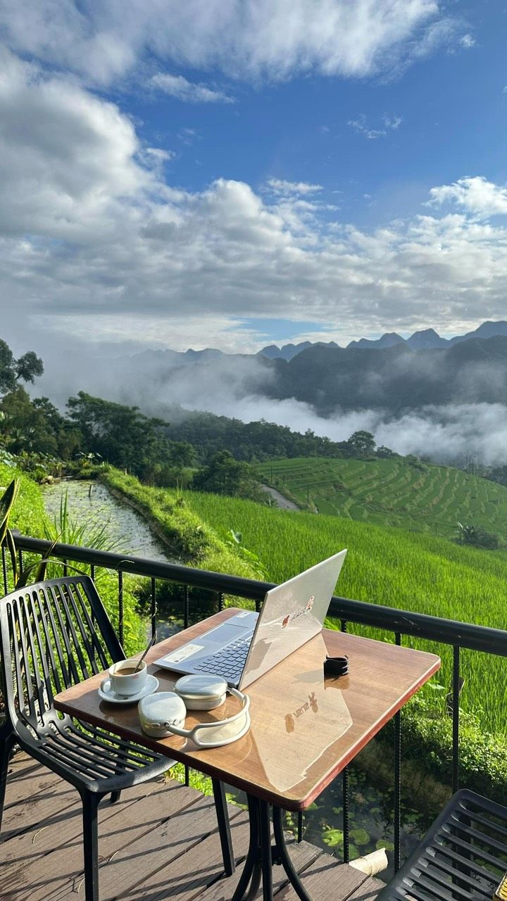 Ett träbord med en bärbar dator, en kopp kaffe och en smartphone på en balkong med utsikt över terrasserade risfält och ett bergigt landskap.