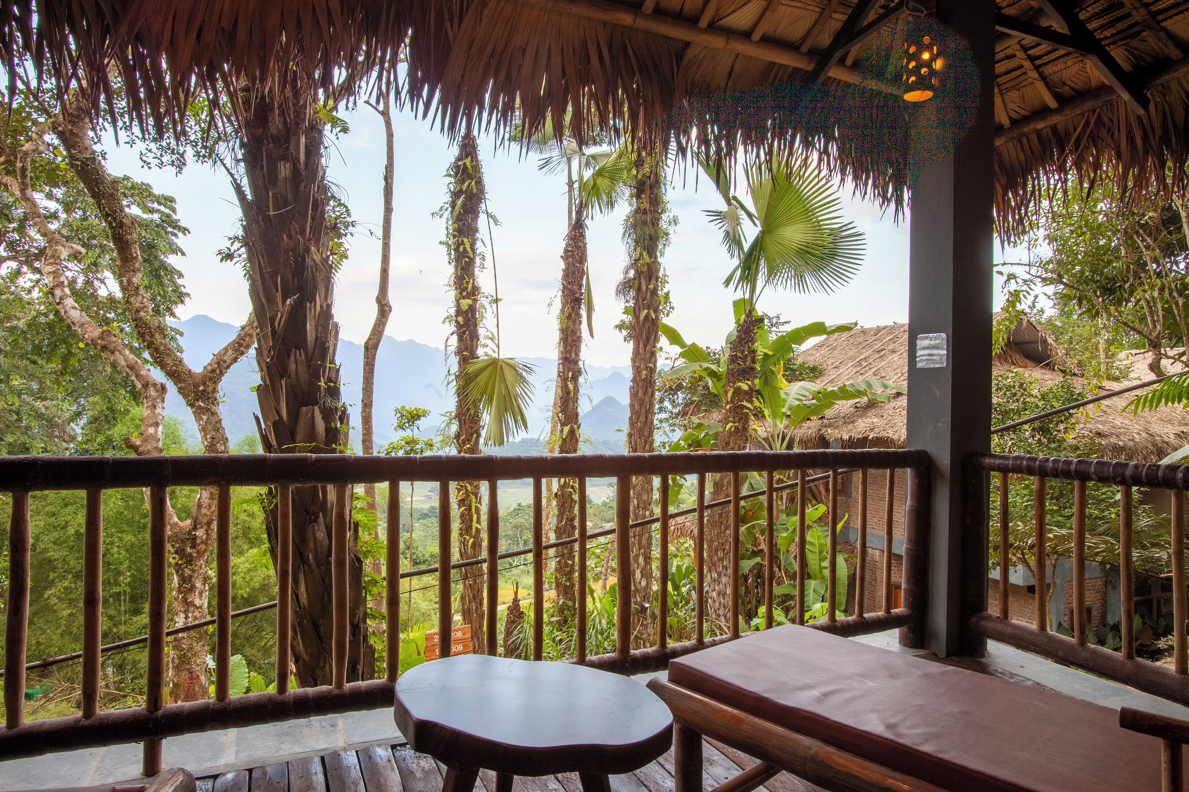 A wooden deck with a thatched roof, overlooking a lush landscape with trees and mountains.