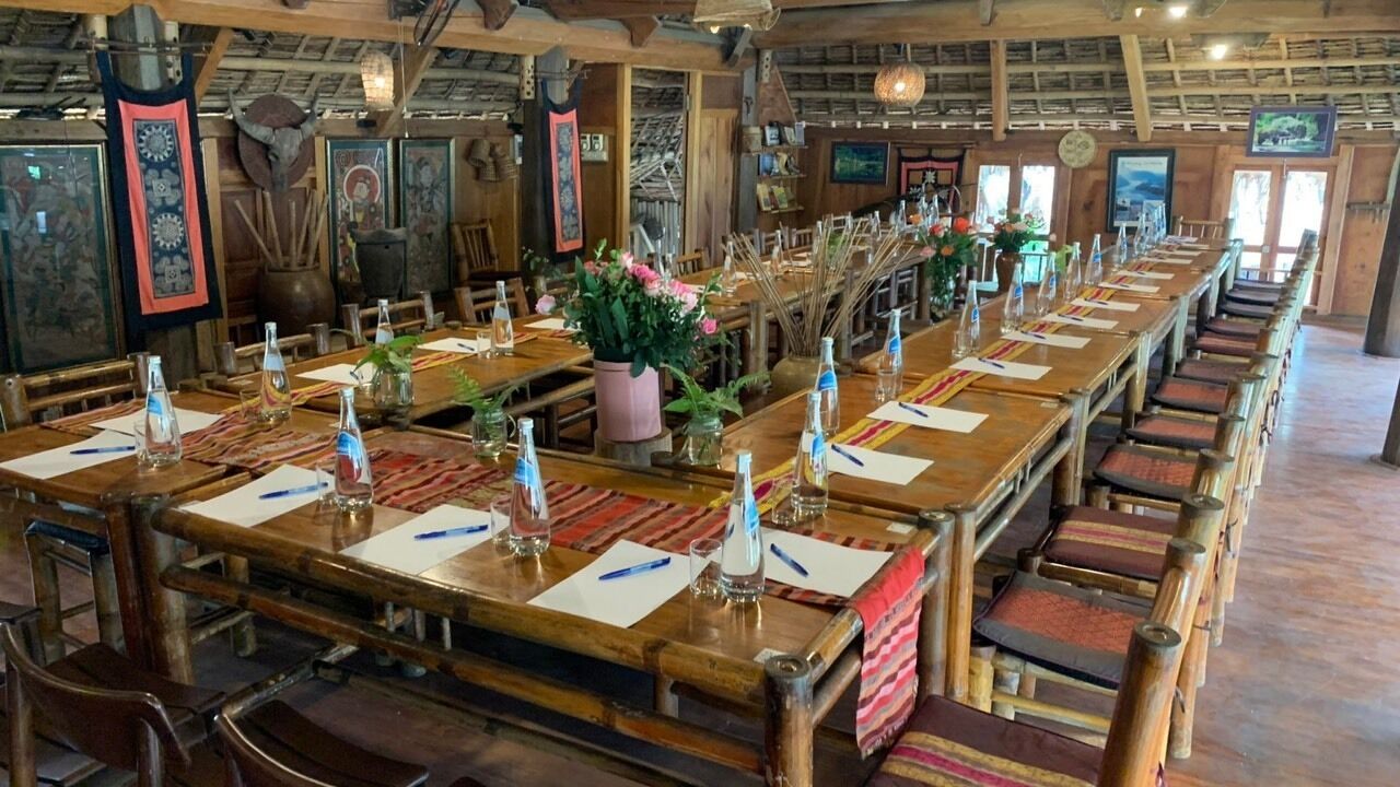 Una larga mesa de comedor de madera con sillas, preparada con copas y servilletas, en un interior rústico con vigas de madera y obras de arte en las paredes.