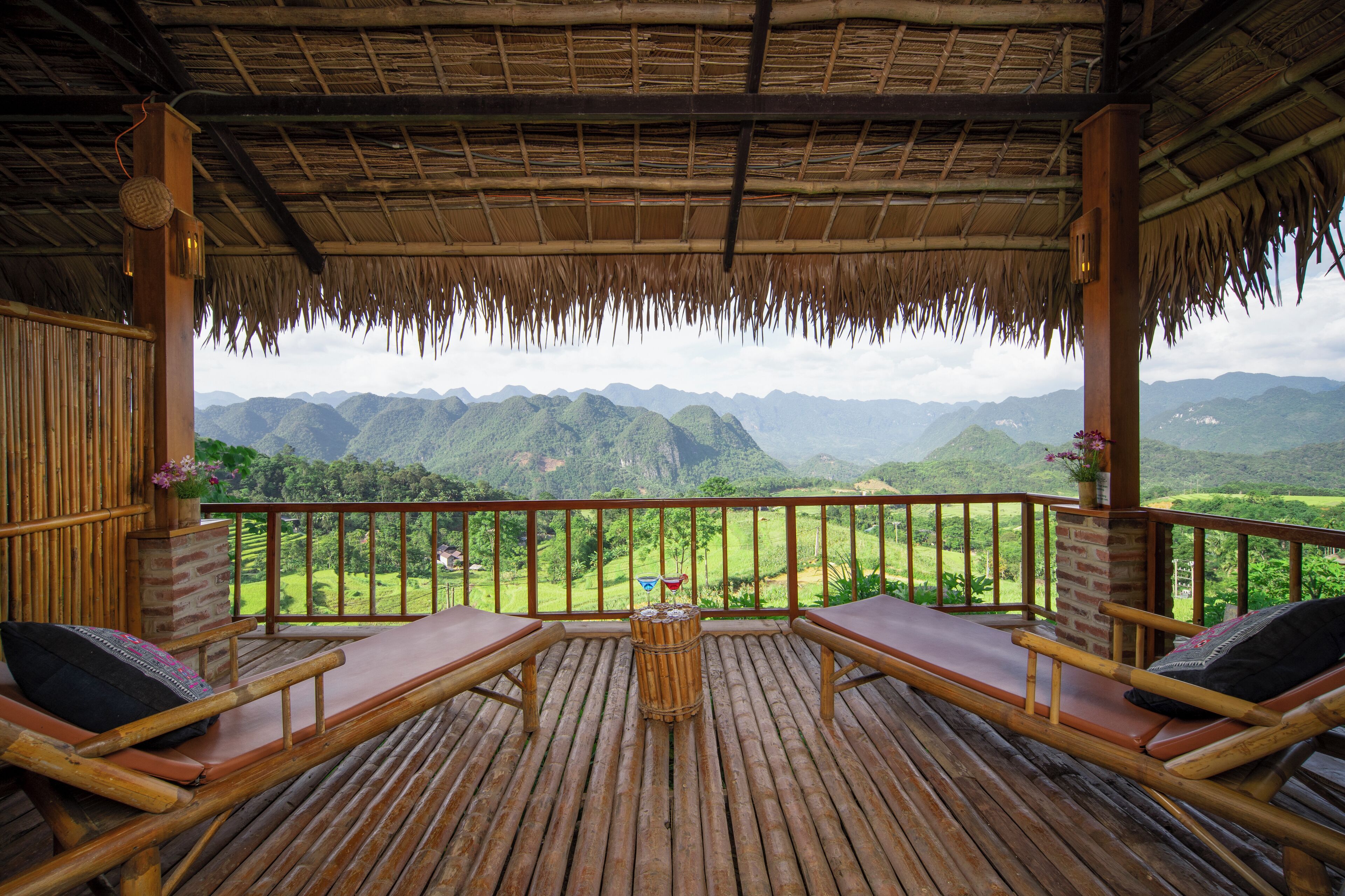 A wooden deck with two cushioned chairs and a small table, overlooking a lush valley with mountains in the distance.