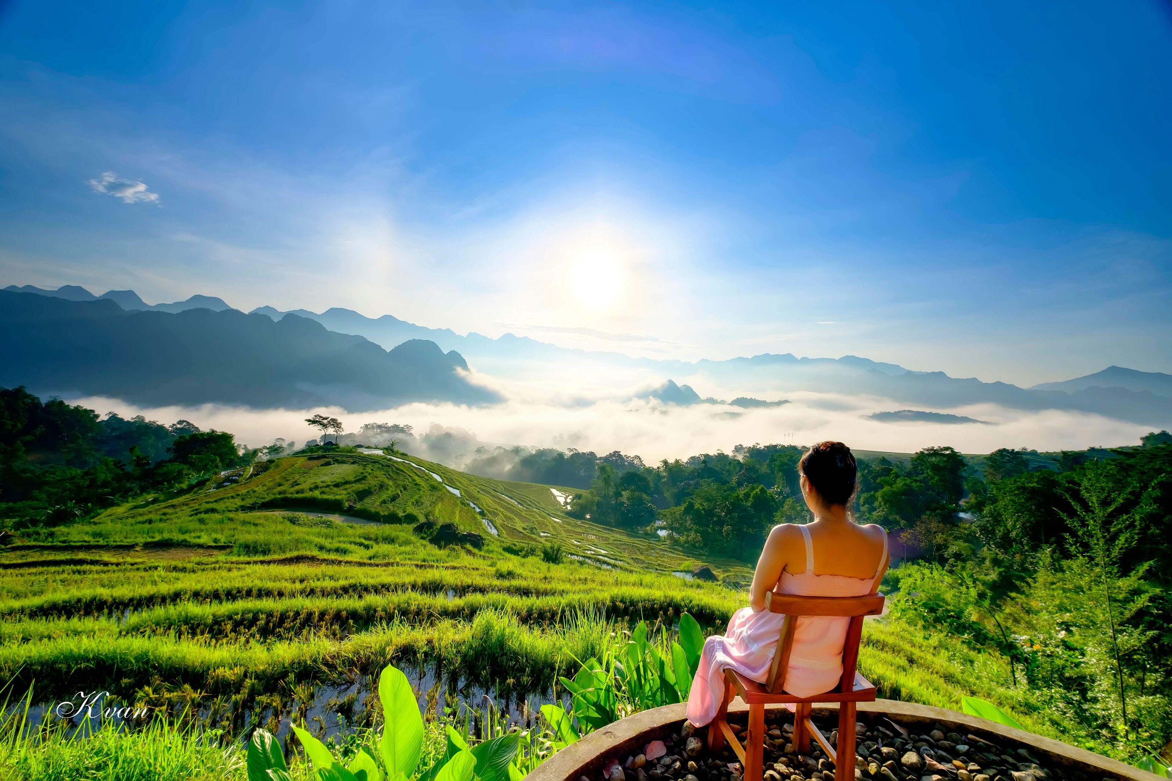 A person sitting on a chair overlooking a lush landscape with terraced fields and mountains in the distance.