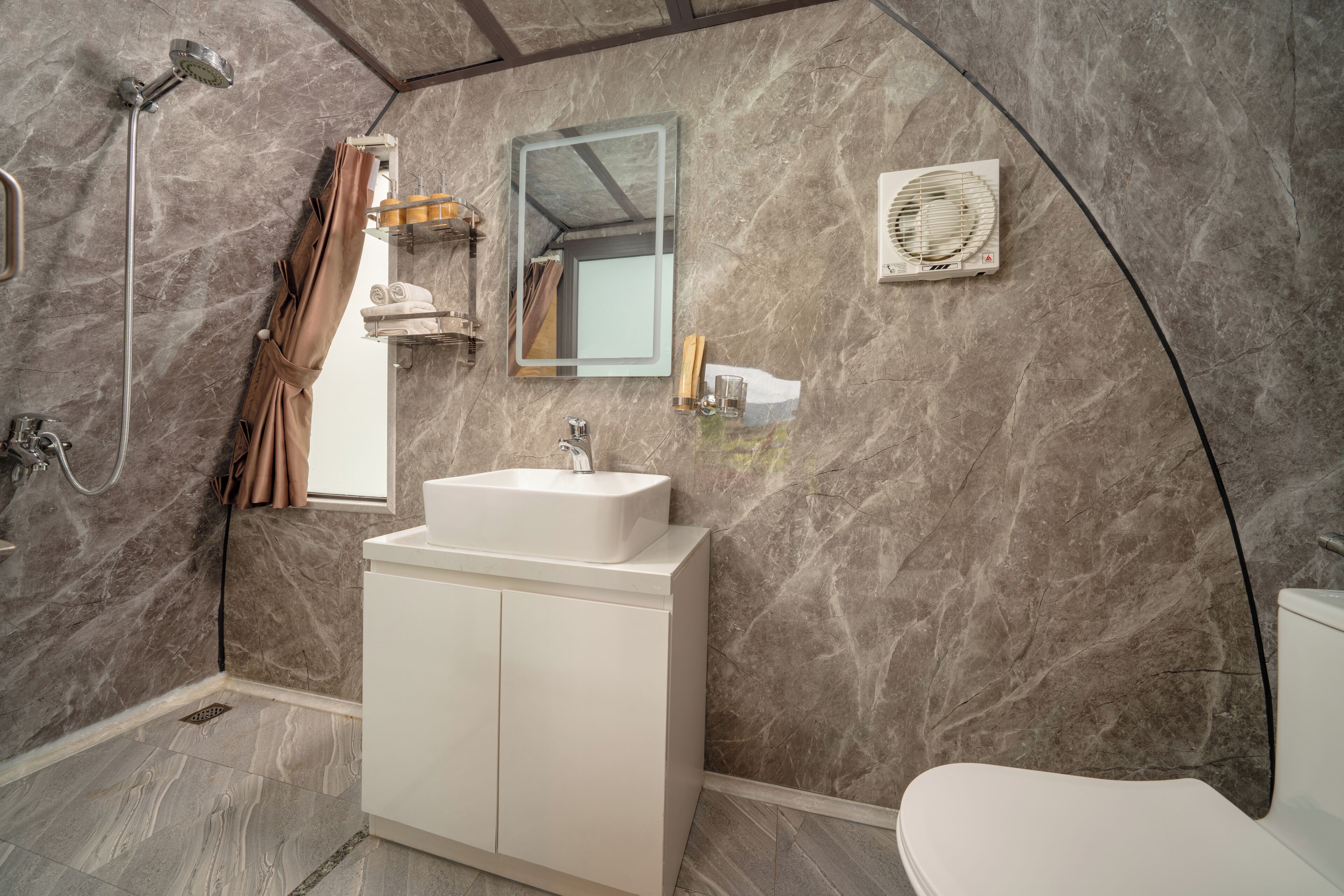 A bathroom with a marble wall, a white sink, a mirror, a shower, and a toilet.