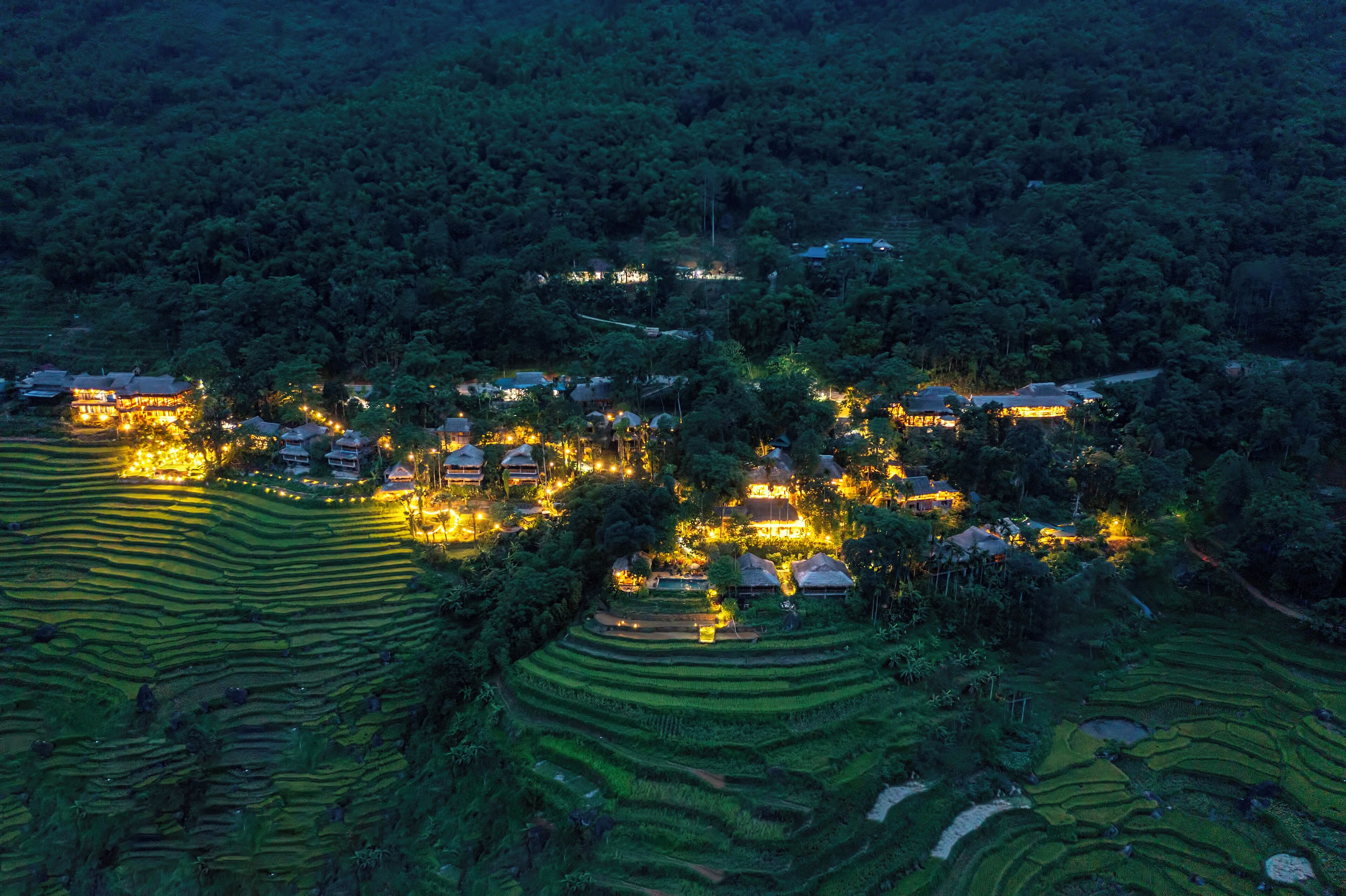 Un champ de rizières en terrasses, avec des bâtiments illuminés, probablement un complexe hôtelier ou un hôtel, entouré d’une forêt dense.