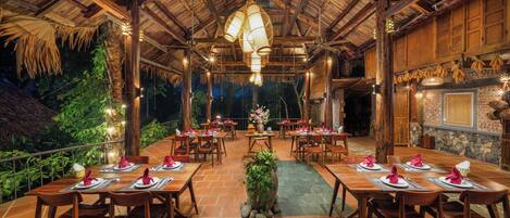 An outdoor dining area with wooden tables and chairs, a thatched roof, and hanging light fixtures.