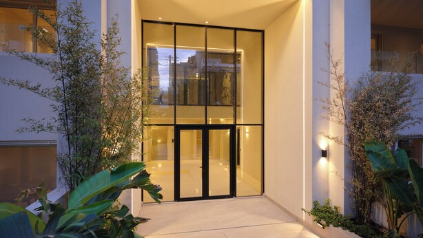 A modern building entrance with glass doors, a lighted pathway, and surrounding greenery. - The Samirah Luxury Residence C4 (Elliniko - Argiroupoli)