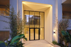 A modern building entrance with glass doors, a lighted pathway, and surrounding greenery. - The Samirah Luxury Residence C4 (Elliniko - Argiroupoli)