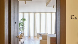 A spacious living area with a wooden floor, a sofa, a dining table with chairs, and a large window with curtains. - The Samirah Luxury Residence C4 (Elliniko - Argiroupoli)