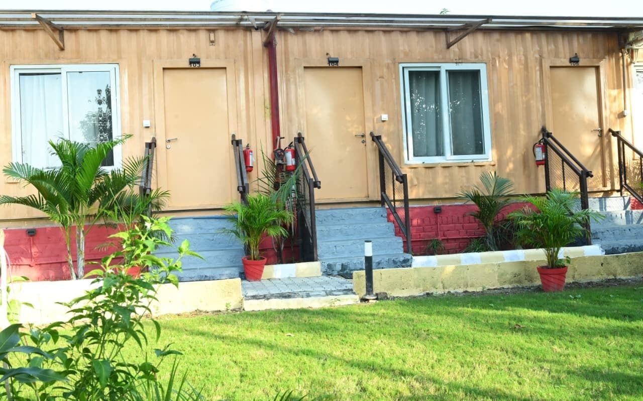 Un bâtiment à deux étages, avec une façade de couleur beige, une base en briques rouges et un petit jardin à l’avant, agrémenté de plantes en pot.