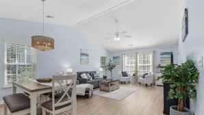 A spacious living area with a dining table, chairs, a sofa, and a coffee table. There are two ceiling fans, large windows with blinds, and a potted plant.