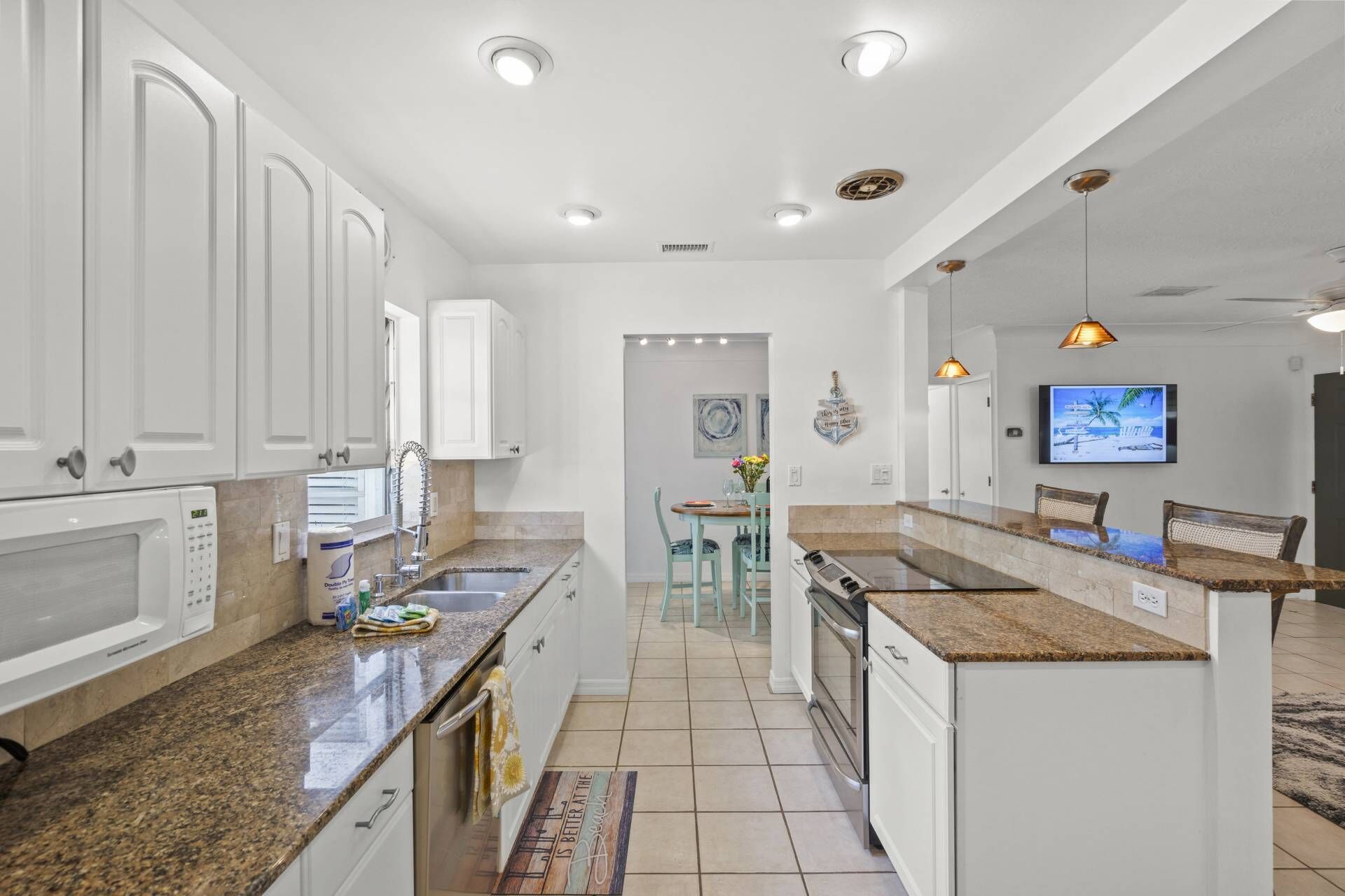 A modern kitchen with white cabinetry, granite countertops, and a central island. There is a dining area with a table and chairs, and a television mounted on the wall.