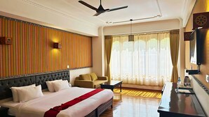 Ein Hotelzimmer mit einem großen Bett, einem Schreibtisch, einem Stuhl, einem Deckenventilator und einem großen Fenster mit Vorhängen.