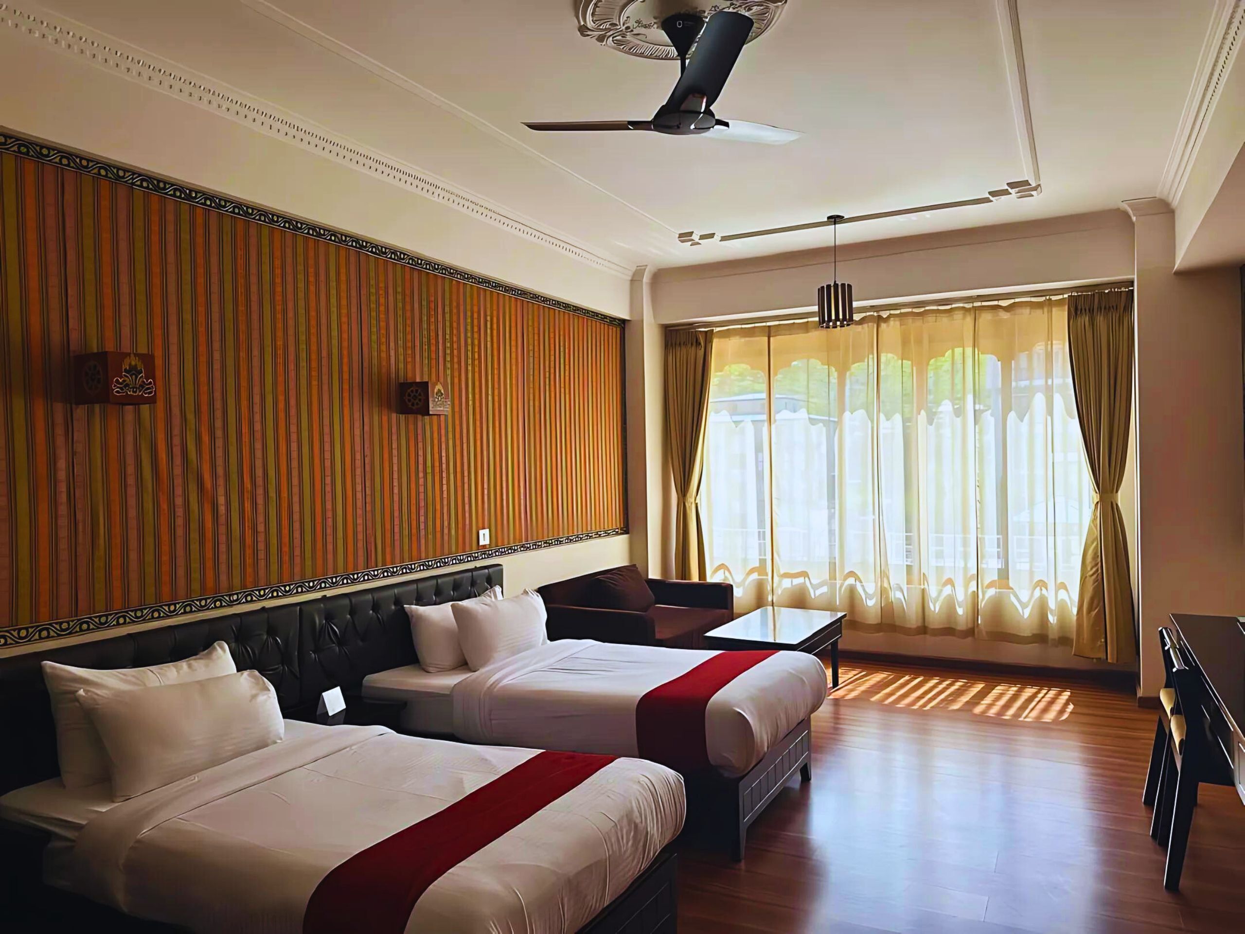 Habitación de hotel con dos camas, un ventilador de techo, un escritorio y un ventanal con cortinas.