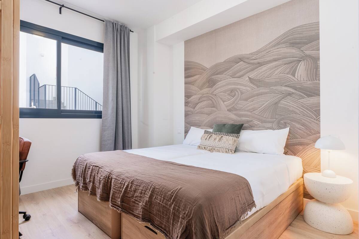 A bedroom with a large bed, a view of a building outside, and a wall with a wave pattern.