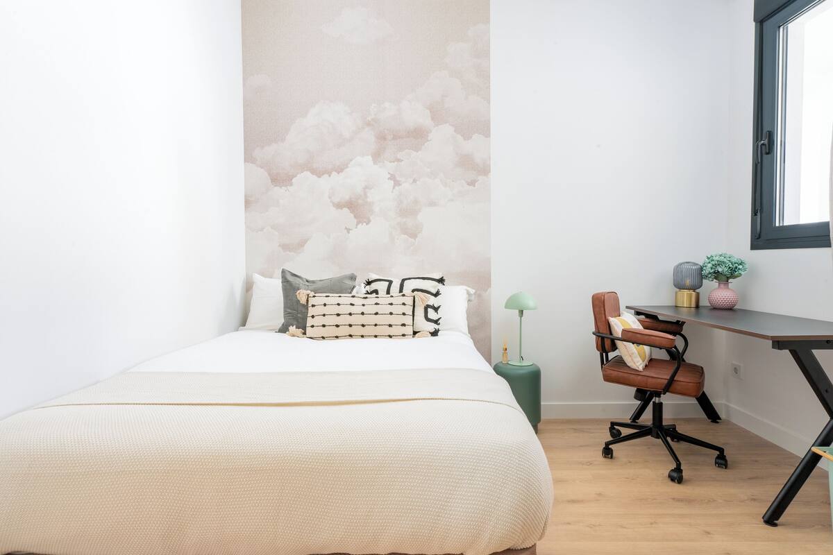 A bedroom with a bed, a desk, a chair, a lamp, and a wall with a cloud mural.