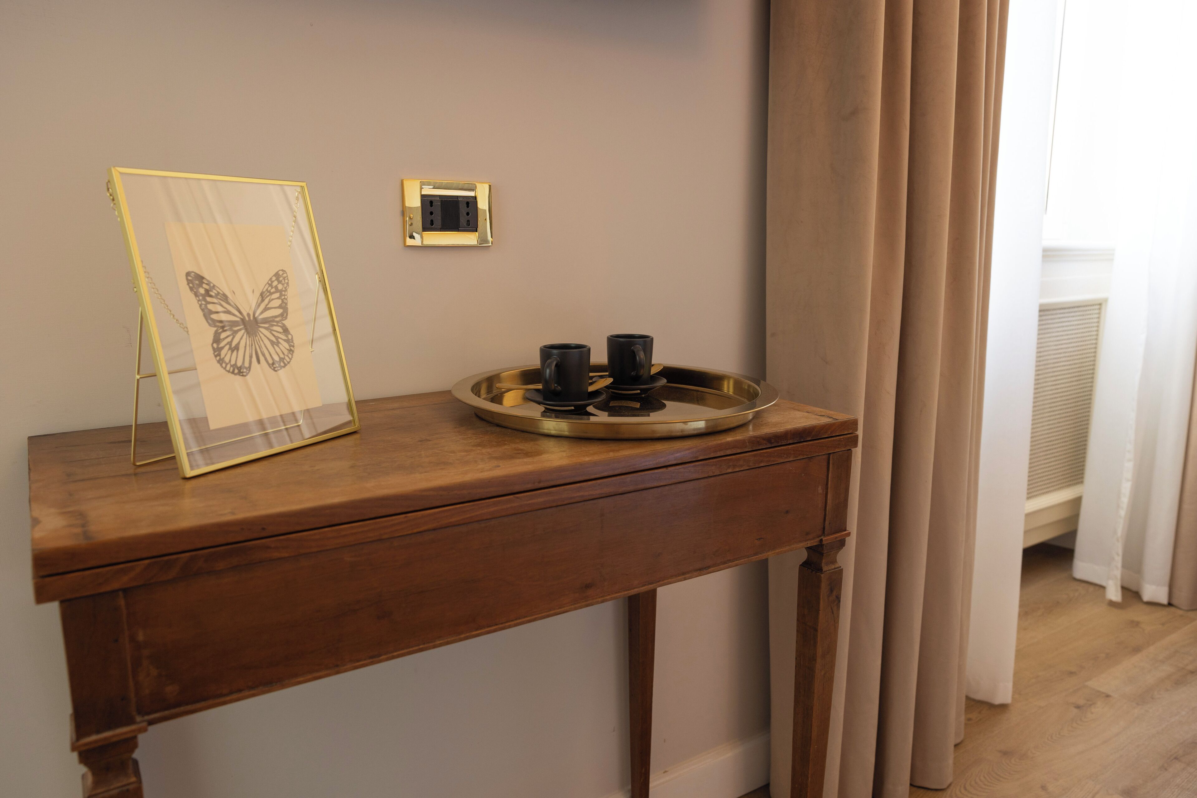 A wooden desk with a framed butterfly artwork, a tray with two black mugs, and a power outlet on the wall.