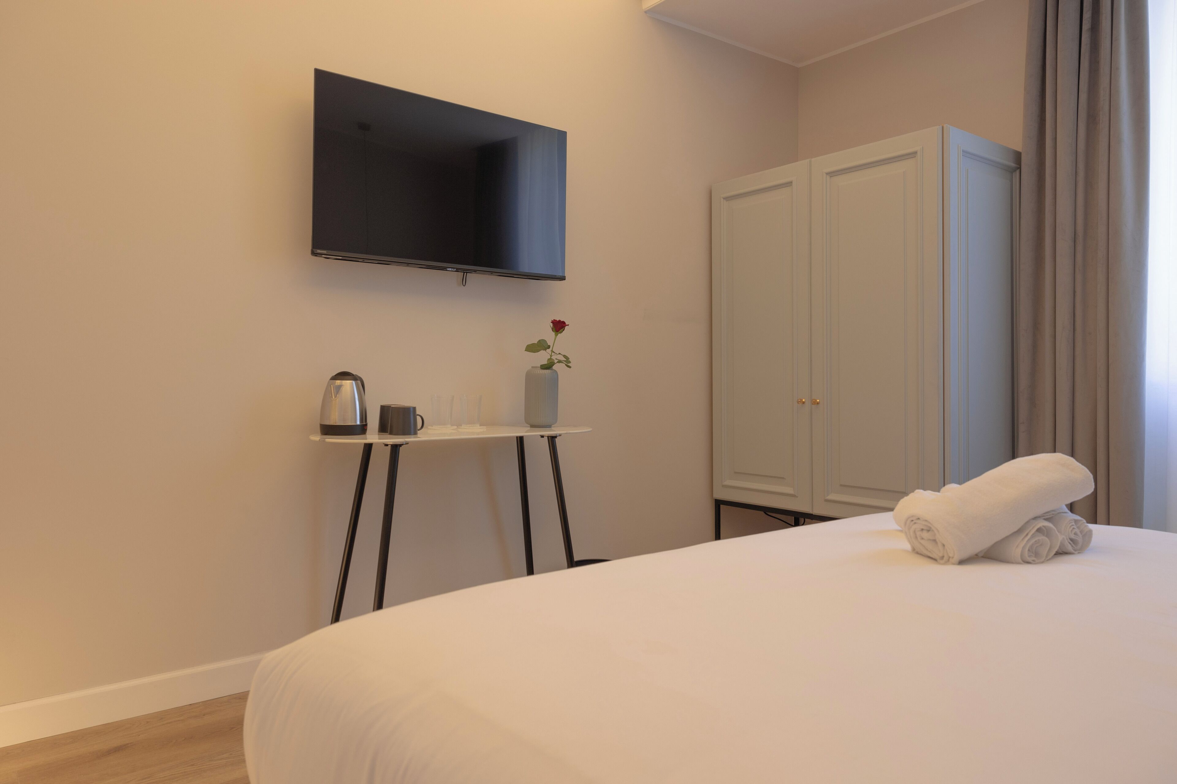 Une chambre d’hôtel avec un lit, un téléviseur fixé au mur, une petite table avec un vase et une tasse, et une armoire.