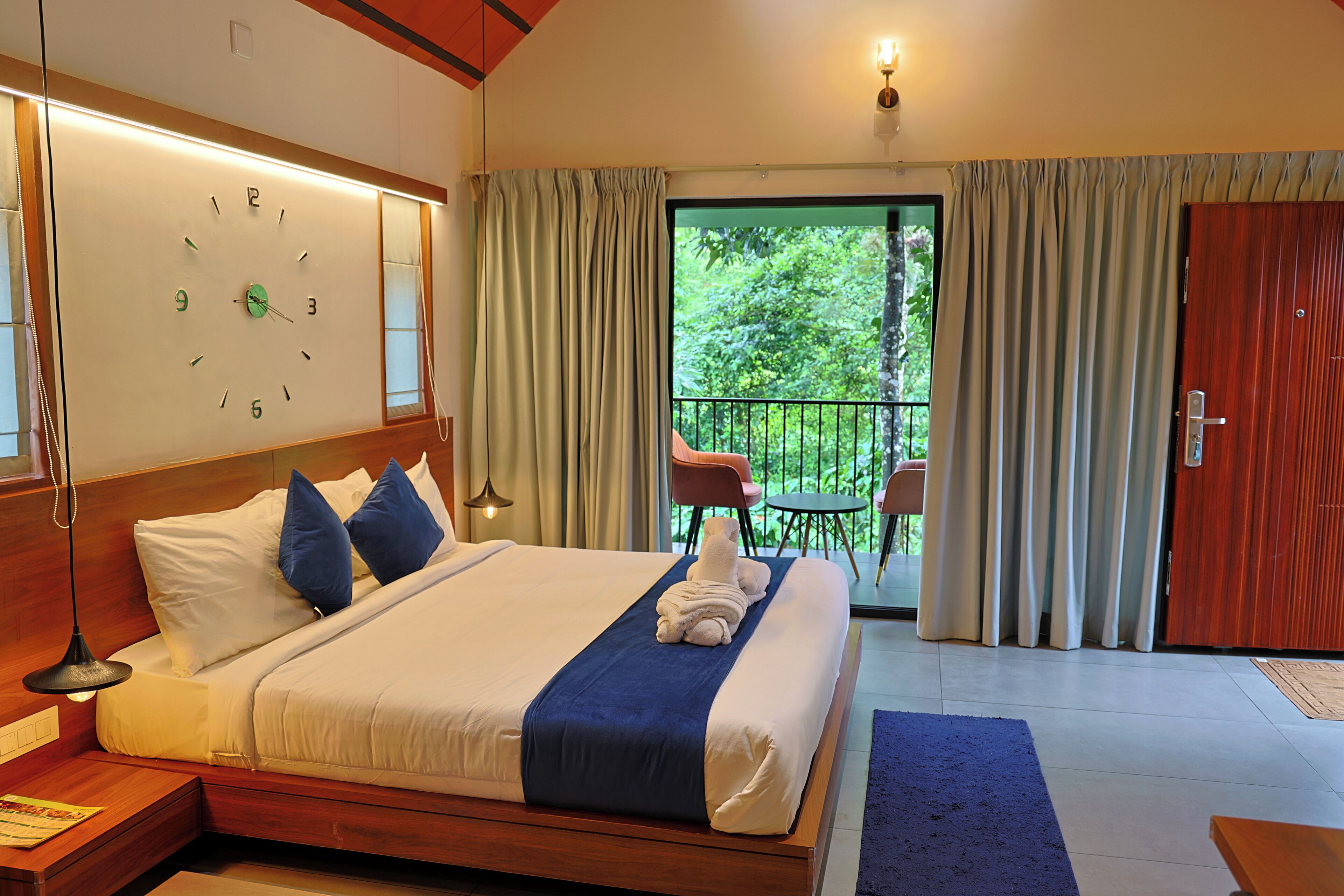 A hotel room with a large bed, a clock on the wall, a balcony with a table and chairs, and a wooden door.