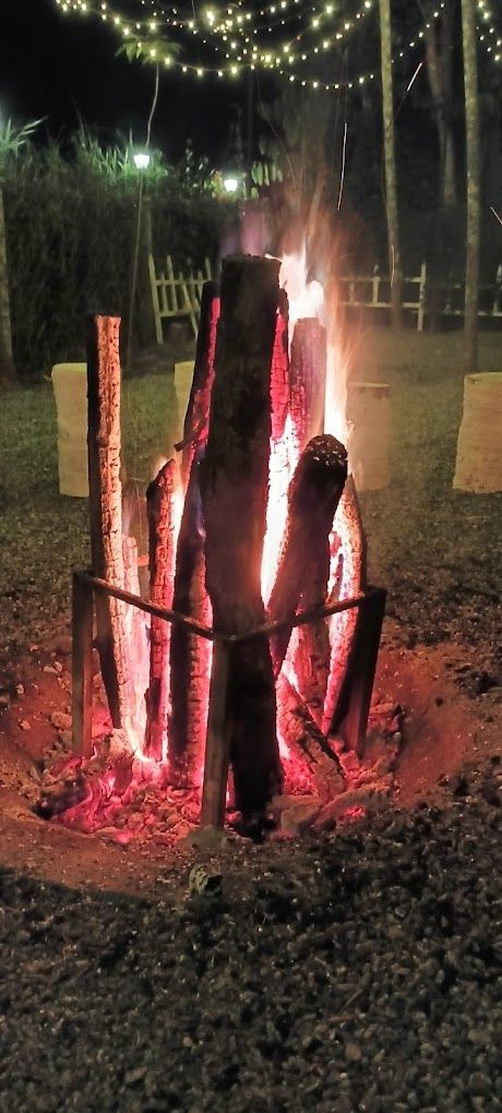 Un feu de camp avec des bûches disposées en forme de tipi, entouré d’une clôture et illuminé par des guirlandes lumineuses en arrière-plan.