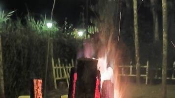 Un feu de camp avec des bûches disposées en forme de tipi, entouré d’une clôture et illuminé par des guirlandes lumineuses en arrière-plan.