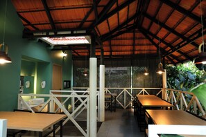 A restaurant with wooden tables and chairs, a high ceiling with exposed beams, and a view of greenery outside.