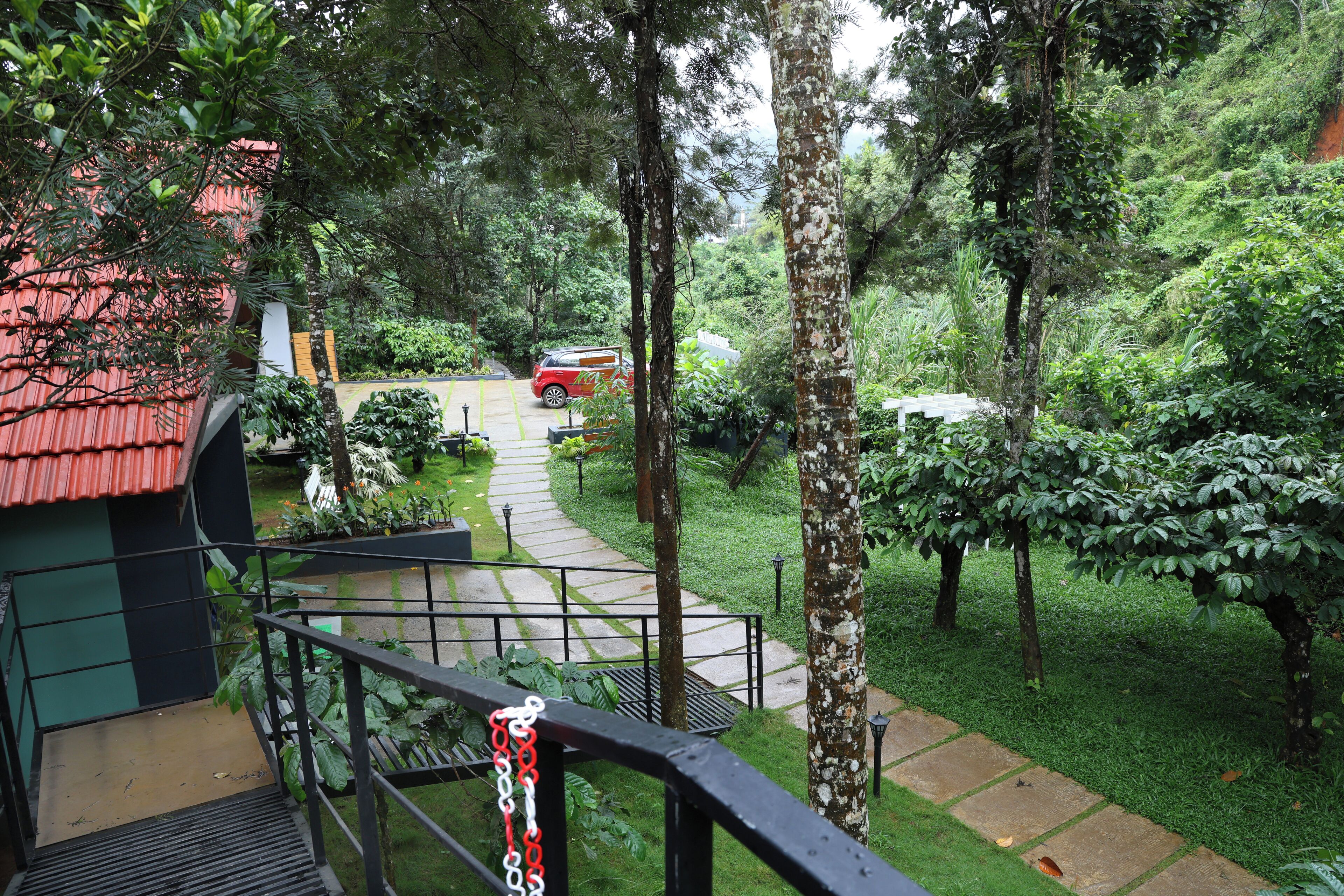 Un jardin luxuriant avec un sentier menant à un véhicule rouge, entouré de verdure et d’un bâtiment doté d’un toit rouge.