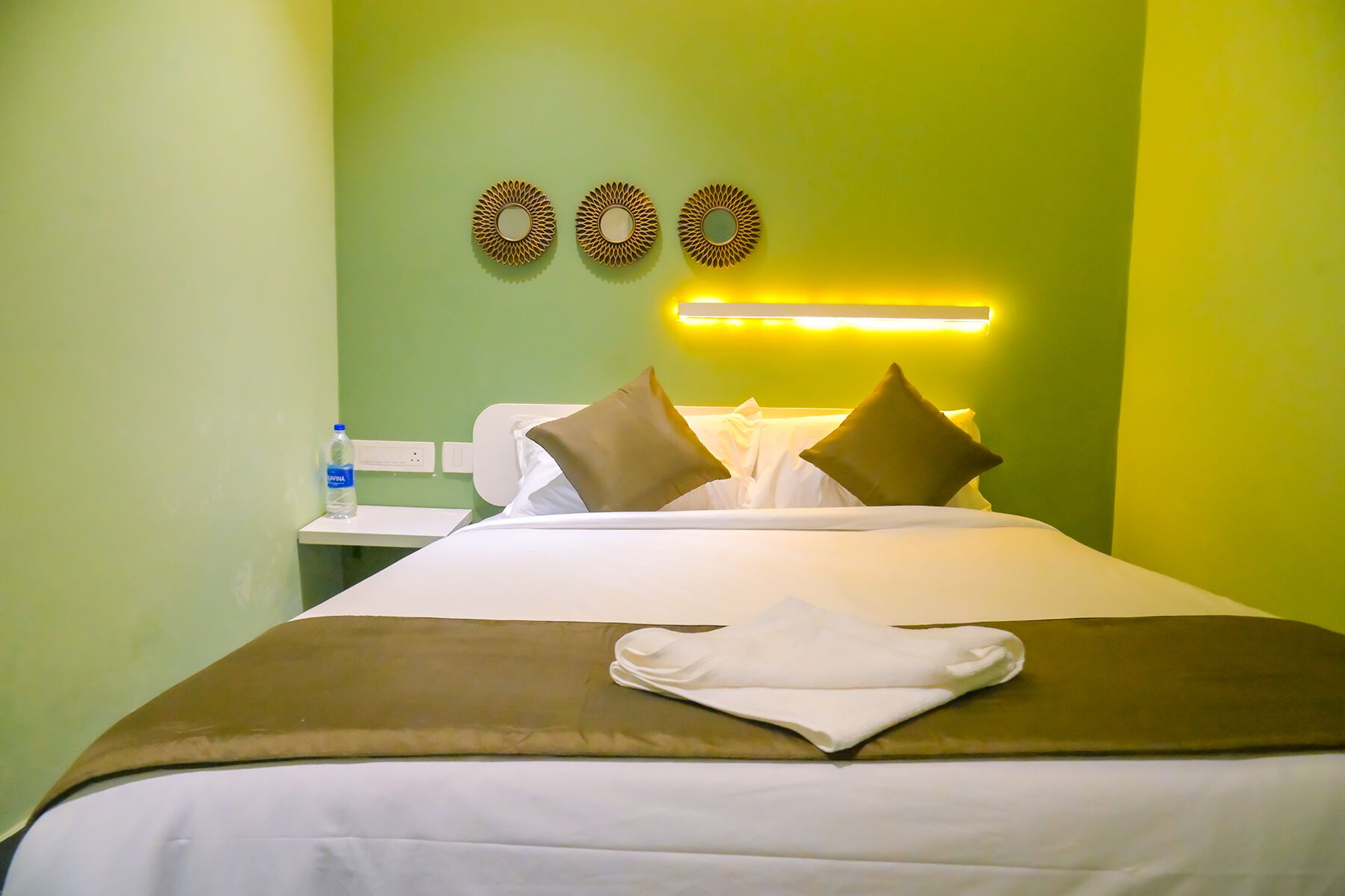 A hotel room with a bed, pillows, a folded towel, a nightstand, a water bottle, and wall decor.